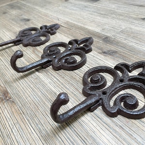 Pode incluir: Três ganchos de metal ornamentados com acabamento marrom escuro. Cada gancho apresenta um design decorativo de rolagem e um gancho curvo.