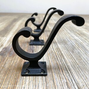 Könnte beinhalten: Vier schwarze Metallhaken mit einem dekorativen, geschwungenen Design. Die Haken sind auf einem quadratischen Sockel montiert und in einer Reihe auf einer Holzoberfläche angeordnet.