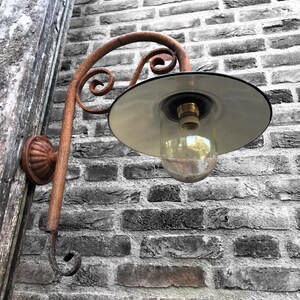 May include: A rusty metal outdoor wall light fixture with a white enamel shade and a clear glass globe. The light is mounted on a brick wall.