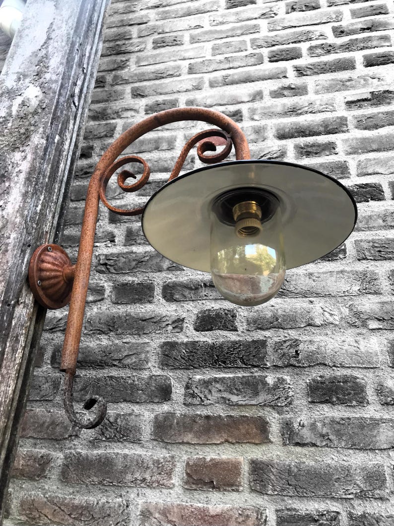 May include: A rusty metal outdoor wall light fixture with a white enamel shade and a clear glass globe. The light is mounted on a brick wall.