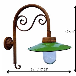 May include: Rustic wall-mounted light fixture with a green metal shade and a clear glass globe. The fixture has a decorative, scrolled metal arm and measures 17.94 inches tall and 17.55 inches wide.