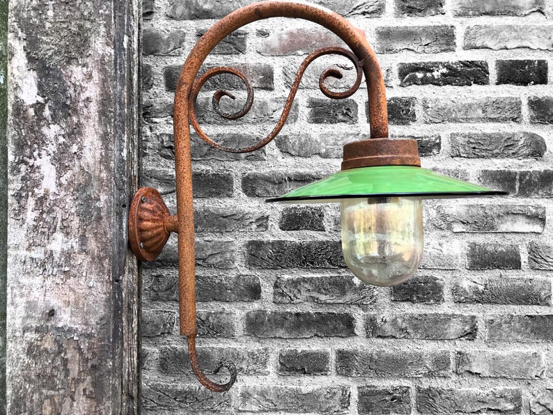 May include: A rusty metal wall-mounted lamp with a green glass shade and a clear glass bulb. The lamp is mounted on a brick wall.