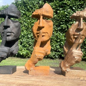 Può includere: Tre sculture astratte di volti con un dito sulle labbra, in nero, arancione e bronzo. Le sculture sono su basi quadrate e sembrano essere realizzate in un materiale strutturato, forse pietra o metallo.