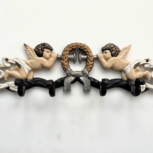 Könnte beinhalten: Ein schwarzer Metall-Kleiderhaken mit zwei Cherubfiguren, die einen Kranz mit Schleife halten. Die Cherubim sind in Beige- und Goldtönen bemalt, und der Kranz ist goldfarben mit einer silbernen Schleife.