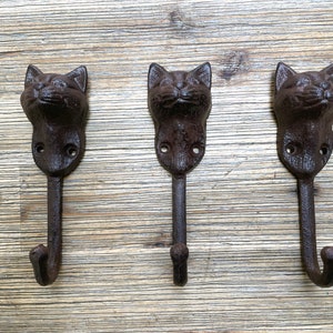 Può includere: Tre ganci a testa di gatto in ghisa marrone montati su una superficie in legno.