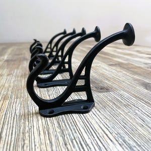 Könnte beinhalten: Sechs schwarze Metallhaken mit dekorativem Design. Die Haken sind an einer Holzoberfläche montiert.