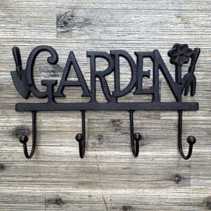 May include: Black metal garden hook rack with three hooks and the word "GARDEN" in decorative lettering. The rack features a small flower and gardening tools on either side of the word.