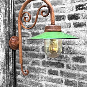 May include: A rusty metal wall-mounted light fixture with a green glass shade and a decorative scroll design. The light fixture is mounted on a brick wall.