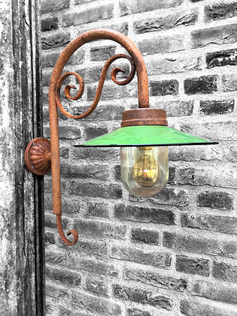 May include: A rusty metal wall-mounted light fixture with a green glass shade and a decorative scroll design. The light fixture is mounted on a brick wall.