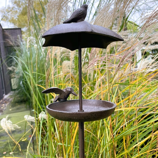 May include: A black metal bird feeder with a small umbrella on top and a bird perched on the umbrella. The feeder has a small dish for food and is designed to be placed in a garden.