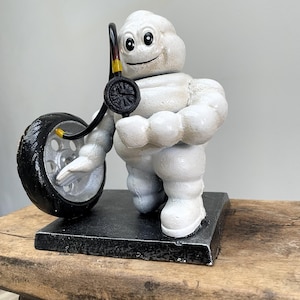 Può includere: Statuetta bianca dell'omino Michelin che tiene in mano uno pneumatico nero e un manometro. La figura è su una base rettangolare nera. Lo pneumatico ha un cerchio argentato. Il manometro ha dettagli gialli. Altezza: circa 15 cm.