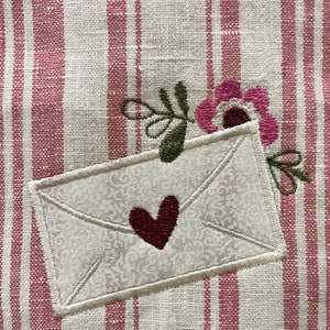 Può includere: Una busta bianca con un cuore rosso ricamato su di essa. La busta è su uno sfondo di tessuto a righe rosa e bianco con un fiore rosa ricamato sul lato destro.