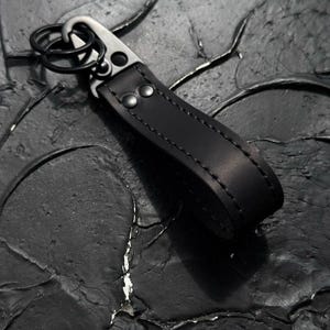 May include: A black leather keychain featuring a matte black metal carabiner clip and a circular split ring. The leather strap is folded into a loop and secured with two matching black metal rivets, with visible black stitching along the edges.