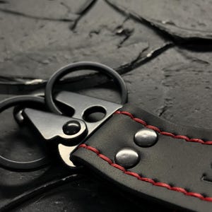 Puede incluir: Primer plano de un llavero de cuero negro con costuras rojas. El llavero presenta un cierre met&aacute;lico y dos remaches met&aacute;licos. Las anillas met&aacute;licas est&aacute;n unidas a la correa de cuero.