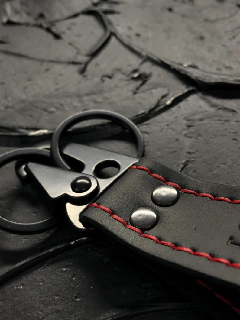 Puede incluir: Primer plano de un llavero de cuero negro con costuras rojas. El llavero presenta un cierre met&aacute;lico y dos remaches met&aacute;licos. Las anillas met&aacute;licas est&aacute;n unidas a la correa de cuero.