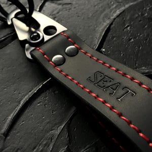 Puede incluir: Llavero de cuero negro con costuras rojas y la palabra "SEAT" grabada. El llavero tiene un cierre de metal plateado y dos remaches. Fondo negro texturizado.
