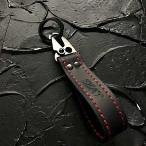 Puede incluir: Llavero de cuero negro con costuras rojas y la palabra "SEAT" en rojo. Cuenta con un cierre de metal plateado y un anillo negro. El fondo es una superficie negra texturizada.