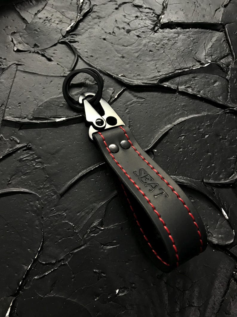 Puede incluir: Llavero de cuero negro con costuras rojas y la palabra "SEAT" en rojo. Cuenta con un cierre de metal plateado y un anillo negro. El fondo es una superficie negra texturizada.