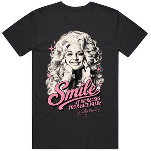 May include: A black short-sleeved t-shirt featuring a black and white portrait of Dolly Parton with voluminous hair. The graphic includes pink star accents and the text "Smile IT INCREASES YOUR FACE VALUE - Dolly Parton" with a small heart symbol.
