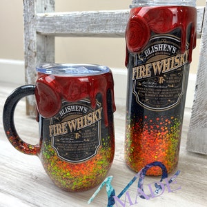 May include: Two stainless steel tumblers with a black and orange glitter finish. The tumblers are decorated with a red wax-like drip and a label that reads "Blishen's Fire Whisky".