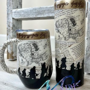 May include: Two black and white ceramic mugs with a map of Middle-earth from The Lord of the Rings printed on them. The mugs have a gold rim and a gold inscription that reads "One Ring to rule them all." The mugs are filled with a black liquid and have a silhouette of the Fellowship of the Ring on the side.
