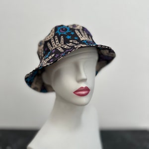Op de afbeelding: Een zwarte bucket hat met een beige en blauwe print van handen, sterren en manen. De hoed zit op een wit mannequin hoofd.