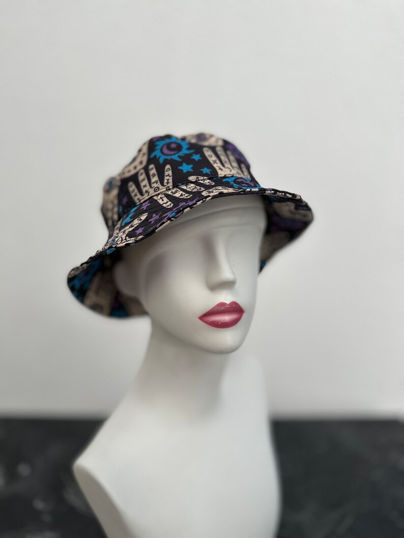 Op de afbeelding: Een zwarte bucket hat met een beige en blauwe print van handen, sterren en manen. De hoed zit op een wit mannequin hoofd.