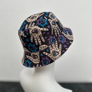 Op de afbeelding: Zwarte bucket hat met een print van handen, ogen, sterren en manen. De print is in tinten blauw, paars en wit op een zwarte achtergrond.
