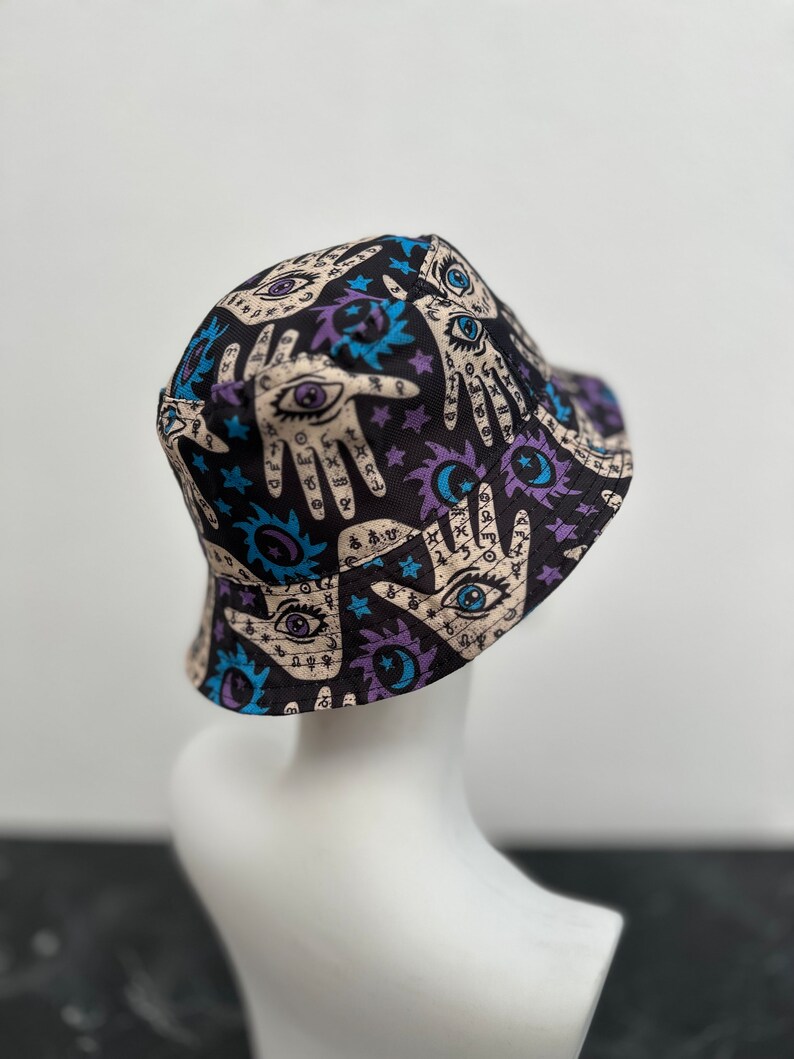 Op de afbeelding: Zwarte bucket hat met een print van handen, ogen, sterren en manen. De print is in tinten blauw, paars en wit op een zwarte achtergrond.