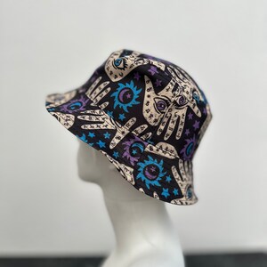 Op de afbeelding: Een zwarte bucket hat met een kleurrijke print van handen, sterren, manen en zonnen. De print heeft een mystiek en occult thema.