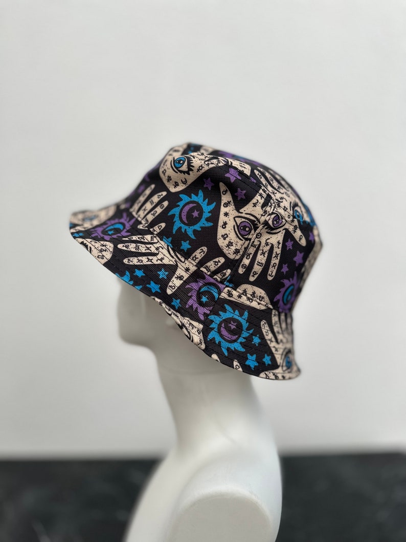 Op de afbeelding: Een zwarte bucket hat met een kleurrijke print van handen, sterren, manen en zonnen. De print heeft een mystiek en occult thema.
