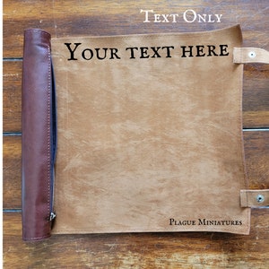 May include: A brown leather gaming mat with a rolled-up edge and two straps. The mat has the text "YOUR TEXT HERE" printed on it. The mat is made by Plague Miniatures.