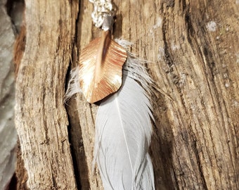 May include: A silver chain necklace with a pendant featuring a gray feather and a copper-colored metal leaf. The necklace is displayed on a piece of weathered wood.