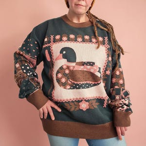 May include: A dark green long-sleeve knit sweater featuring a central cream-colored square panel with a mallard duck graphic. The design includes pink floral borders, brown ribbed cuffs and hem, and patchwork-style patterns on the sleeves.