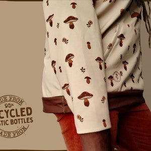 May include: A cream-colored hoodie with a brown mushroom print. The hoodie has a brown drawstring and a brown pocket. The hoodie is made from 95% recycled plastic bottles. The text "MADE FROM 95% RECYCLED PLASTIC BOTTLES MADE FROM" is printed on a brown circle.