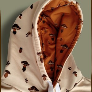 May include: A beige hoodie with a brown mushroom print. The hood is lined with a brown fabric with a smaller mushroom print. The hoodie has a white drawstring.