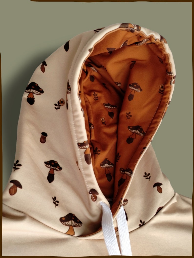 May include: A beige hoodie with a brown mushroom print. The hood is lined with a brown fabric with a smaller mushroom print. The hoodie has a white drawstring.