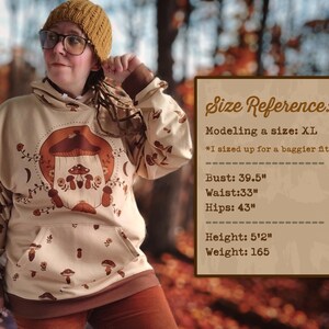 May include: A beige hoodie with a mushroom graphic and the text "Shroom Queens". The model is wearing a size XL and is 5'2" tall and weighs 165 pounds.