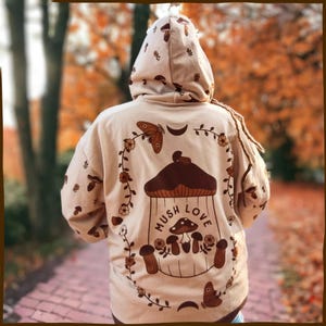 May include: A beige hoodie with a brown mushroom graphic and the text "Mush Love". The graphic features a mushroom with a house-like top and a crescent moon. There are also butterflies and flowers on the hoodie.