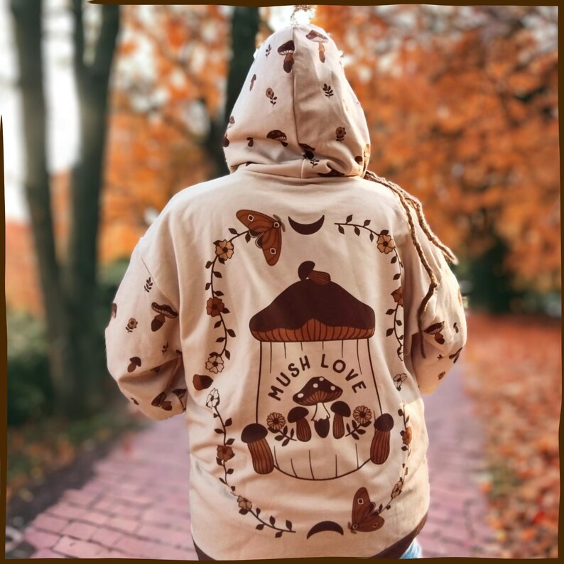 May include: A beige hoodie with a brown mushroom graphic and the text "Mush Love". The graphic features a mushroom with a house-like top and a crescent moon. There are also butterflies and flowers on the hoodie.