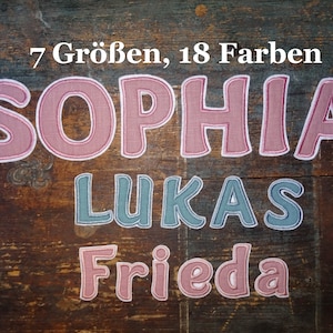 Könnte beinhalten: Drei Stoffaufnäher mit den Namen SOPHIA, LUKAS und FRIEDA in gestickter Schrift. Der Text 7 Größen, 18 Farben steht oben. Die Aufnäher sind in Altrosa und gedecktem Blau gehalten und verfügen über weiße Umrandungen.