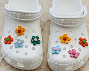 May include: A pair of white clogs with colorful flower charms. The clogs have the word "crocs" printed on the strap.