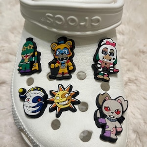 May include: White Crocs shoe with six colorful Jibbitz charms featuring characters from the Five Nights at Freddy's video game franchise. The charms include a green and yellow character with a guitar, a yellow character with a top hat and microphone, a white and red character with a knife, a white and blue character with a jester hat, a yellow sun character with a toothy grin, and a white and purple character with a knife.