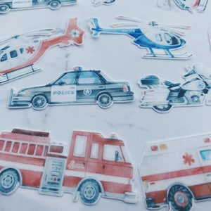 May include: A collection of watercolor-style stickers featuring emergency vehicles. Includes a police car, ambulance, fire truck, police motorcycle, and helicopters. The stickers are in shades of red, blue, white, and gray.