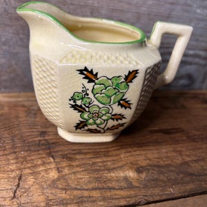 May include: A small, cream-colored ceramic creamer with a green rim and a floral design in shades of green, brown, and yellow.