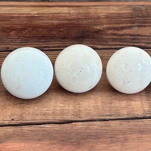 May include: Five round, off-white cabinet knobs with a speckled appearance are arranged in a row on a wooden surface. The knobs have a glossy finish and are evenly spaced, offering a simple, yet elegant design for home decor.