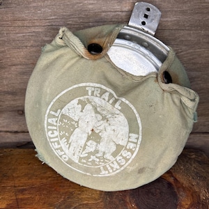 May include: An olive green canteen with a metal cup and a canvas cover. The cover has a white graphic with the words "OFFICIAL TRAIL MESS KIT" in a circular design. The canteen is secured with two black buttons.