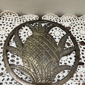 May include: A round brass trivet with a cutout design of a pineapple. The trivet has a smooth, polished finish and is perfect for protecting surfaces from hot dishes.