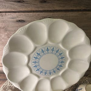 May include: A round, off-white ceramic oyster plate featuring twelve recessed oval wells arranged in a circle. The center of the plate is decorated with a blue pattern of repeating fleur-de-lis symbols enclosed in rectangular frames.