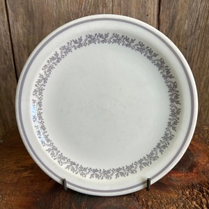 May include: A white ceramic plate with a grey floral design around the rim. The plate is on a wooden surface.